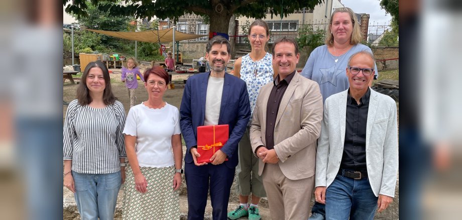 Mit Minister Sven Teuber steht Bürgermeister Thomas Przybylla, Fachbereichsleiter Sven Normann, Kita-Leiterin Tanja Reuter und die drei Kolleginnen des multiprofessionellen Teams im Garten der Kita Bassenheim. 