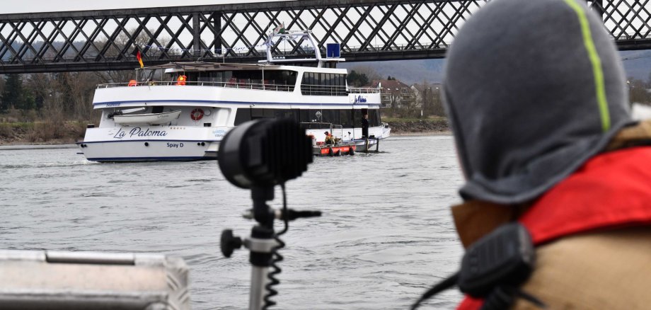 Von einem Feuerwehrboot mit einem Feuerwehrmann im Vordergrund am rechten Bildrand ist das havarierte Schiff und die Eisenbahnbrücke zu sehen.
