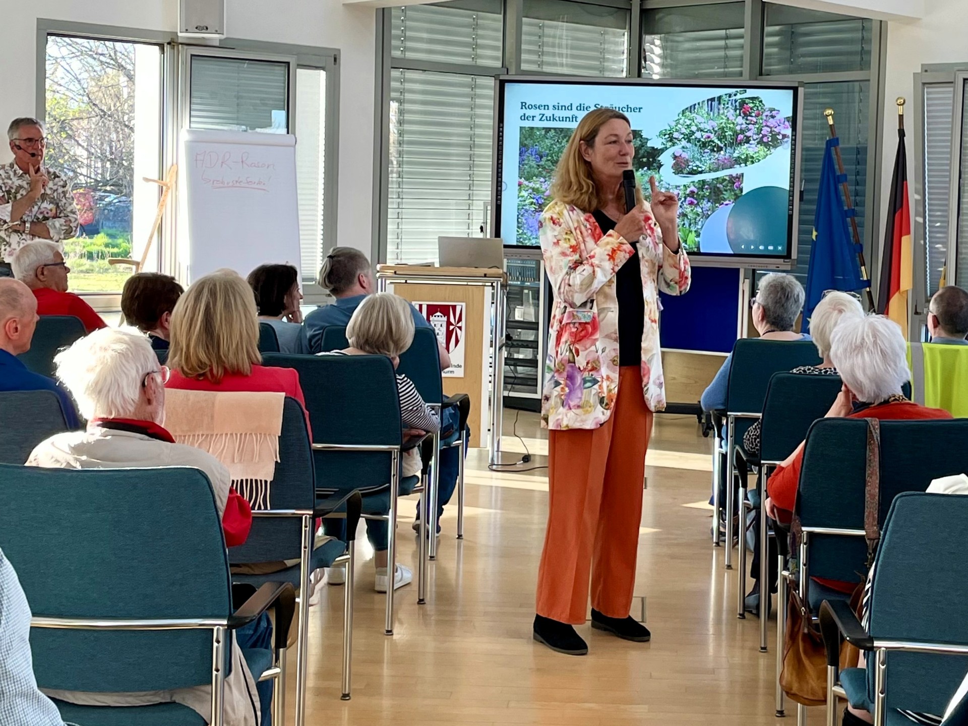 Eröffnung Klimaschutzreihe Referentin Heike Boomgaarden Heike Boomgarden steht mit einem Mikrofon in der Hand in einem Blazer mit Blumenmuster zwischen den beiden Blöcken mit Stuhlreihen, auf denen das Publikum sitzt.