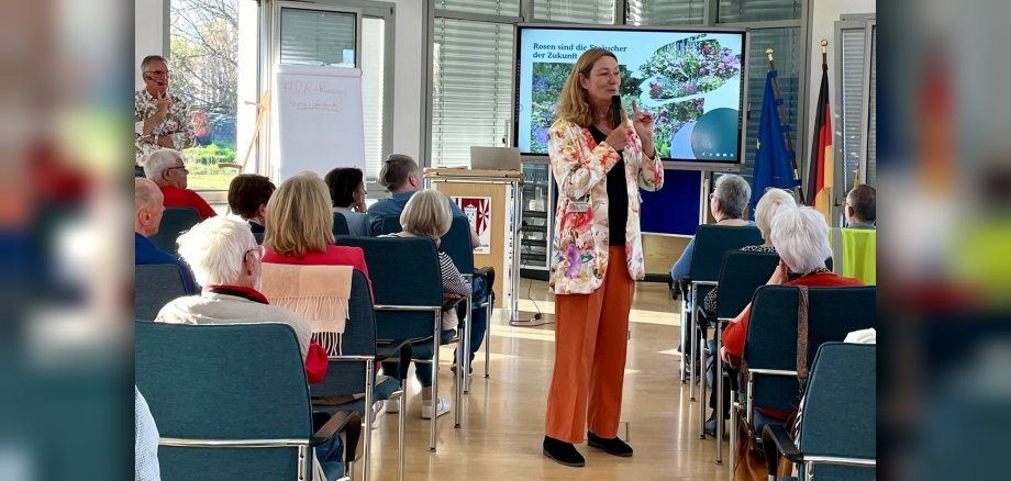 Heike Boomgarden steht mit einem Mikrofon in der Hand in einem Blazer mit Blumenmuster zwischen den beiden Blöcken mit Stuhlreihen, auf denen das Publikum sitzt. 