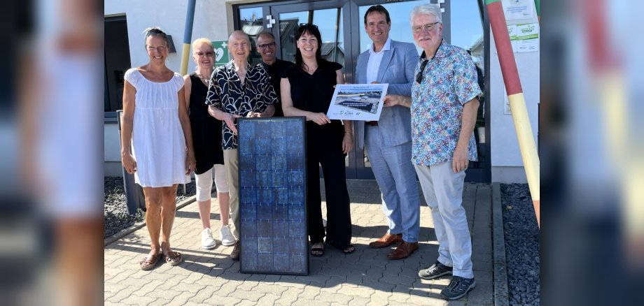 Vor dem Eingang der Kita Schillerstraße stehen  Bürgermeister Thomas Przybylla mit der Kita-Leiterin Kirsten Zimmer und Vertreterinnen und Vertretern der Klimafit-Gruppe, die ein Solarmodul und ein kleines Plakat in der Hand halten. 