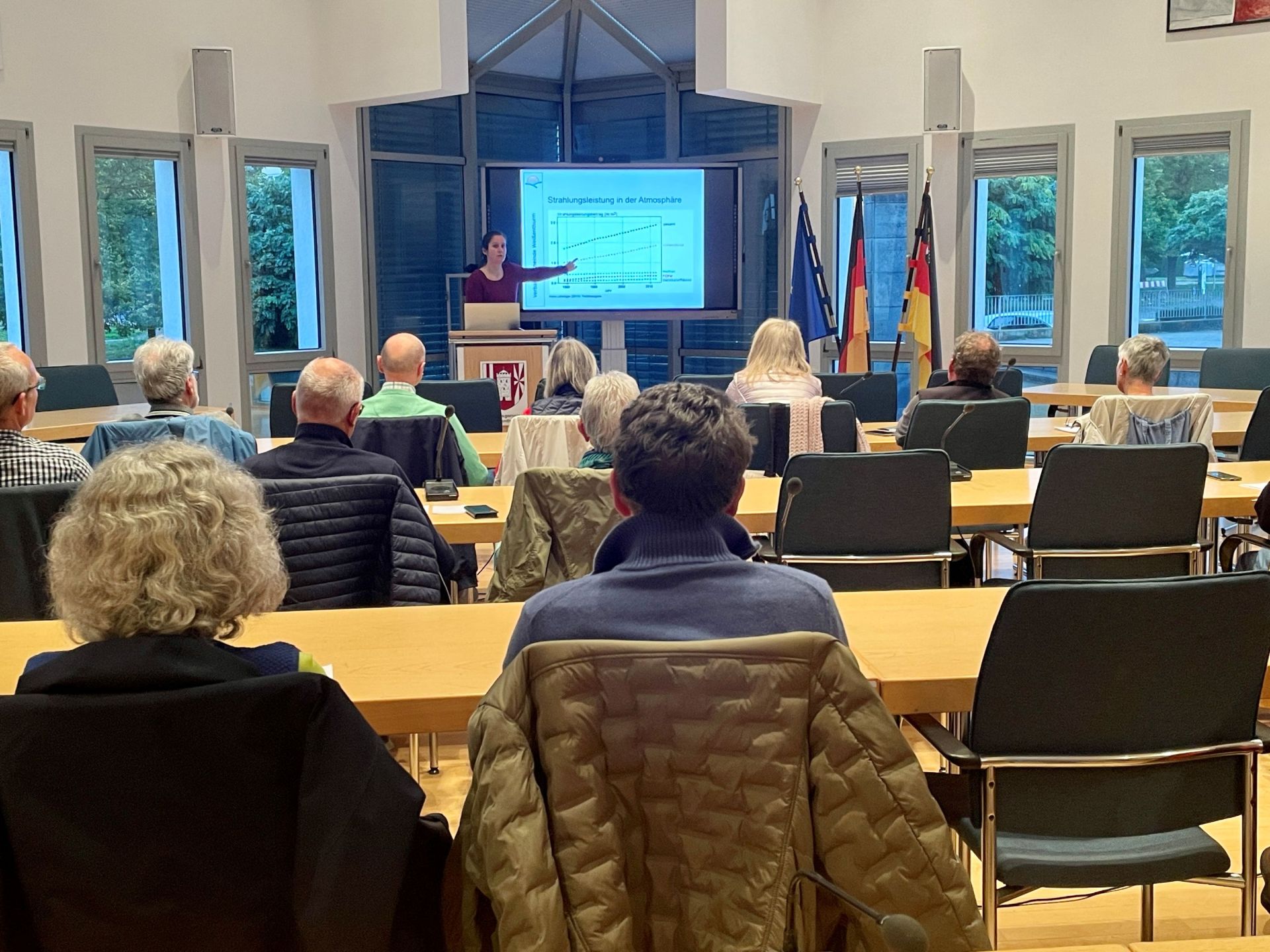 Vortrag Klimawandel 1 Im Ratssaal ist Publikum an Tischen zu sehen vor einem Bildschirm, auf dem die Referentin eine Grafik zeigt.