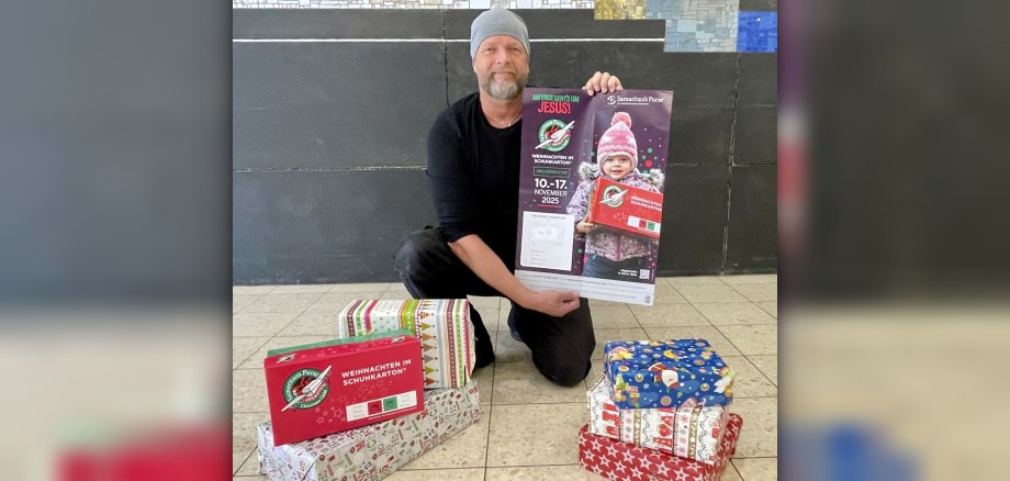 Initiator Uwe Fink zeigt ein Plakat der Aktion Weihanchtei im Schuhkarton, vor ihm stehen weihnachtlich verpackte Geschenke. 