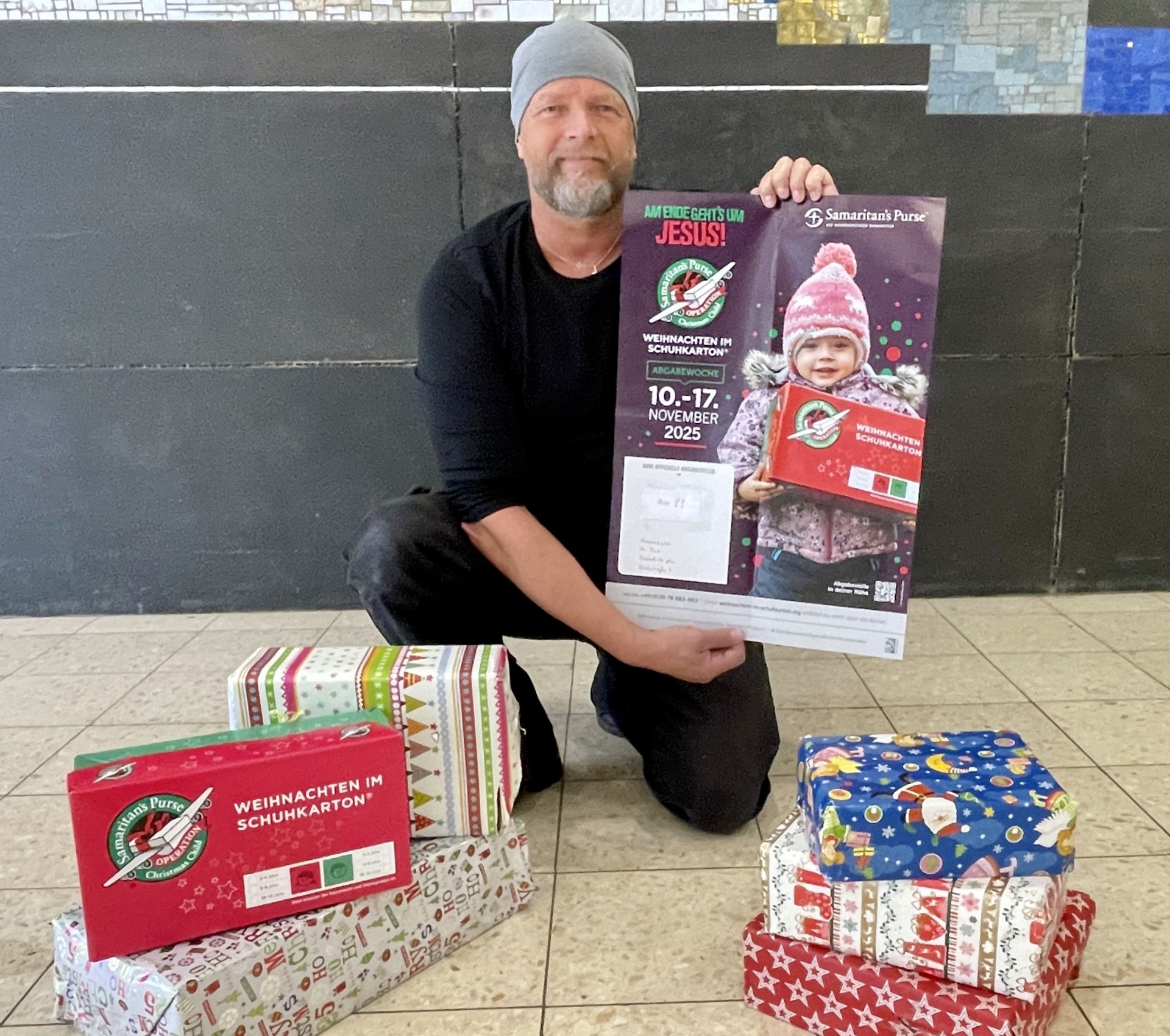 Weihnachten im Schuhkarton Initiator Uwe Fink zeigt ein Plakat der Aktion Weihanchtei im Schuhkarton, vor ihm stehen weihnachtlich verpackte Geschenke.