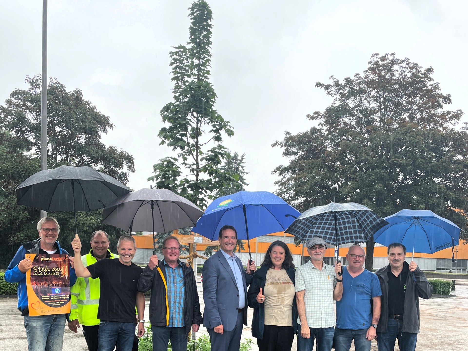 Neue Bäume für das Schulzentrum Unter Regenschirmen stehen vor einem neu gepflanzten Baum Bürgermeister Przybylla, Vertreter der ÖK-Gruppe, des Chores Piano Forte und der anderen, an der Pflanzaktion beteiligten Firmen.