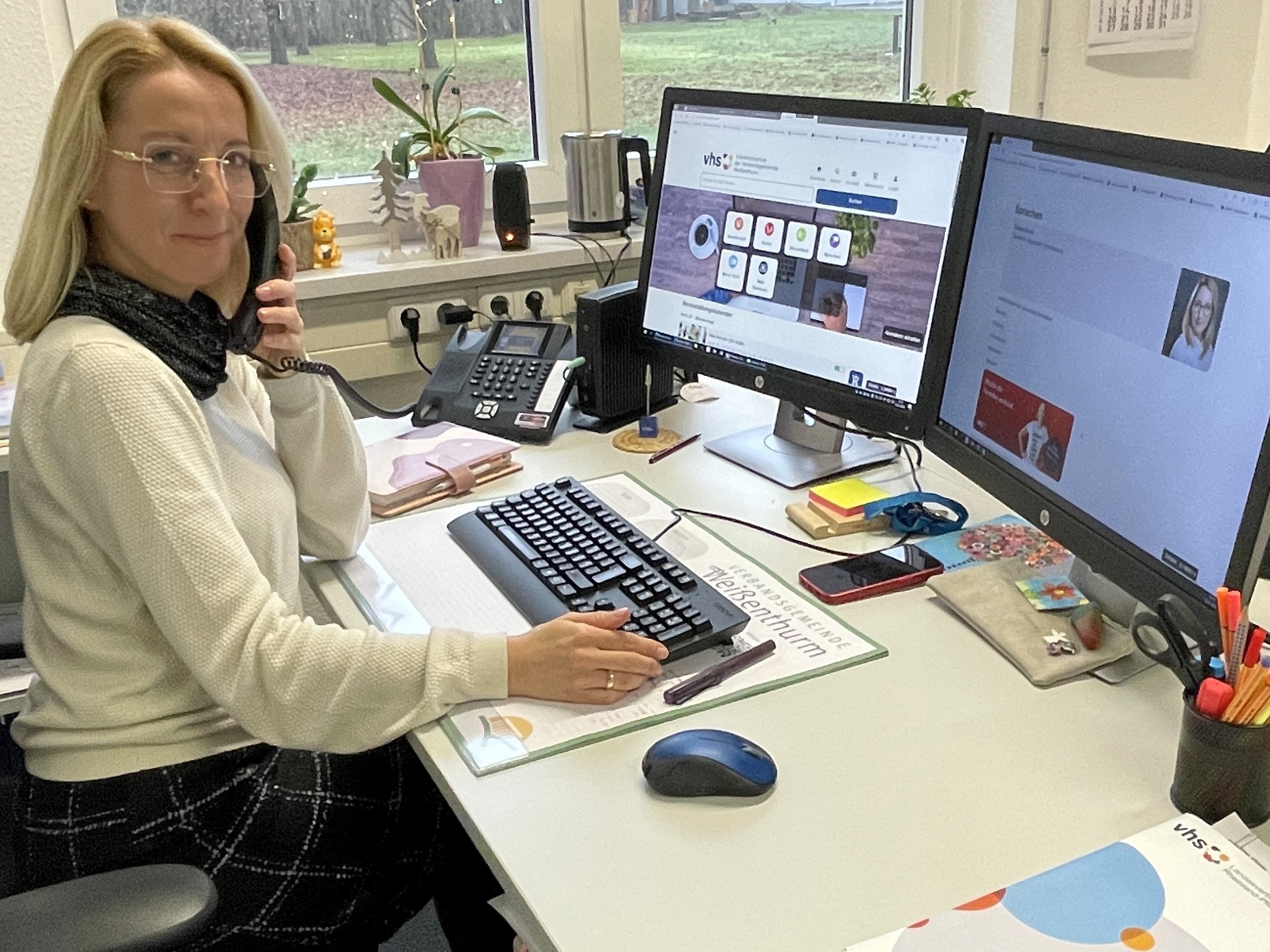 VHS-Mitarbeiterin Suzana Koch sitzt an ihrem Schreibtisch vor zwei Bildschirmen, auf denen die Internetseite der VHS zu sehen ist, und telefoniert.