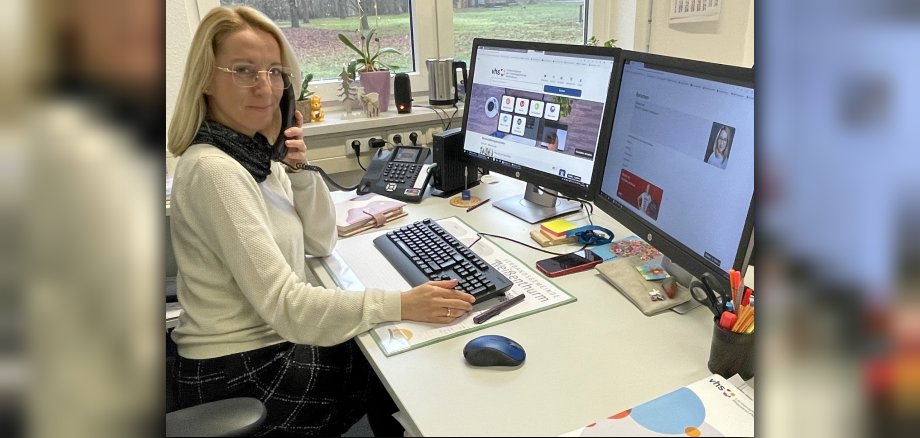 VHS-Mitarbeiterin Suzana Koch sitzt an ihrem Schreibtisch vor zwei Bildschirmen, auf denen die Internetseite der VHS zu sehen ist, und telefoniert.