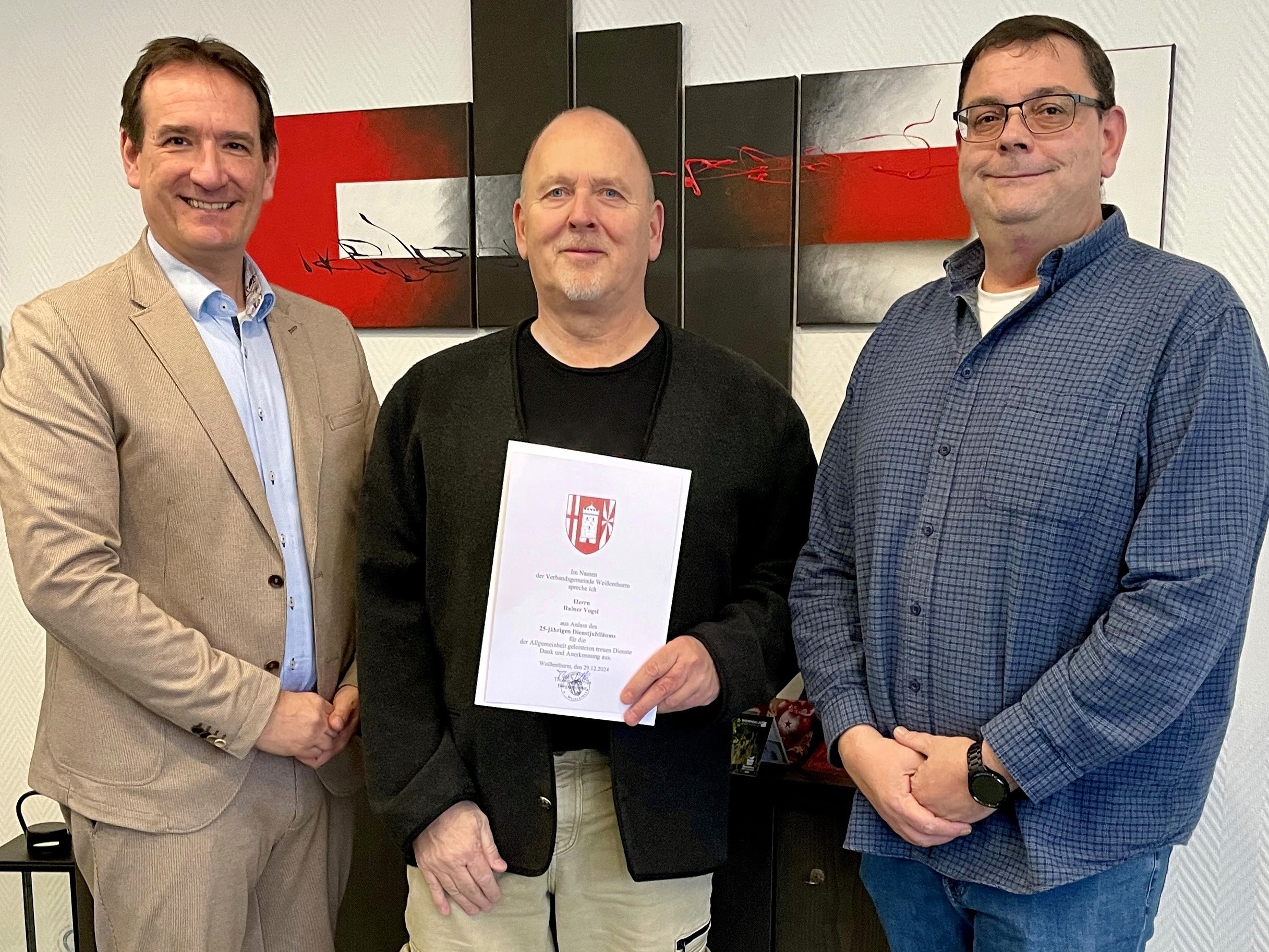 Jubilar Rainer Vogel steht zwischen Bürgermeister Thomas Przybylla und Personalratsmitglied Marko Röser mit seiner Urkunde in der Hand vor einer modernen schwarz-weiß-roten Grafik an der Wand.