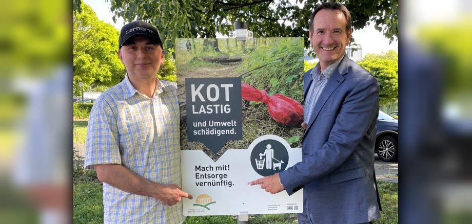Der Initiator der Kampagne, Mesut Özyurt, steht links des Schildes, das dazu aufruft, Hundekot nicht liegenzulassen. Rechts steht Bürgermeister Przybylla.