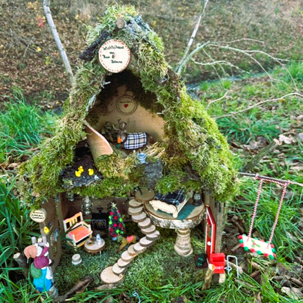 Wichtelweg Ein kleines Wichtelhäuschen mit zwei Stockwerken, einer Wendeltreppe, einem moosbedeckten Dach und kleinen Figuren zwischen Möbeln steht im Wald.