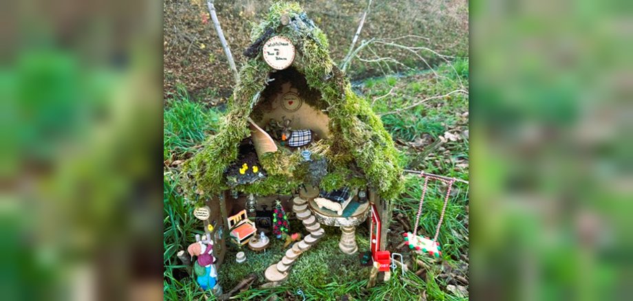 Ein kleines Wichtelhäuschen mit zwei Stockwerken, einer Wendeltreppe, einem moosbedeckten Dach und kleinen Figuren zwischen Möbeln steht im Wald. 
