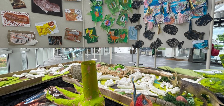 Ausstellungsobjekte sind auf einem Tisch im Vordergrund zu sehen, dahinter Bilder an Wänden. 