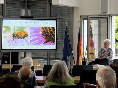 Wildbienenvortrag Foto mit Publikum 1 Referentin Biggi Kaczmarek steht seitlich neben dem Bildschirm mit der Präsentation ihres Vortrags.