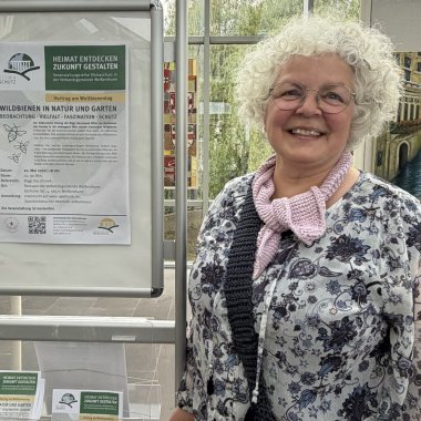 Vortrag Weltbienentag Referentin Referentin Biggi Kaczmarek steht neben einem Info-Ständer, der auf ihren Vortrag hinweist.