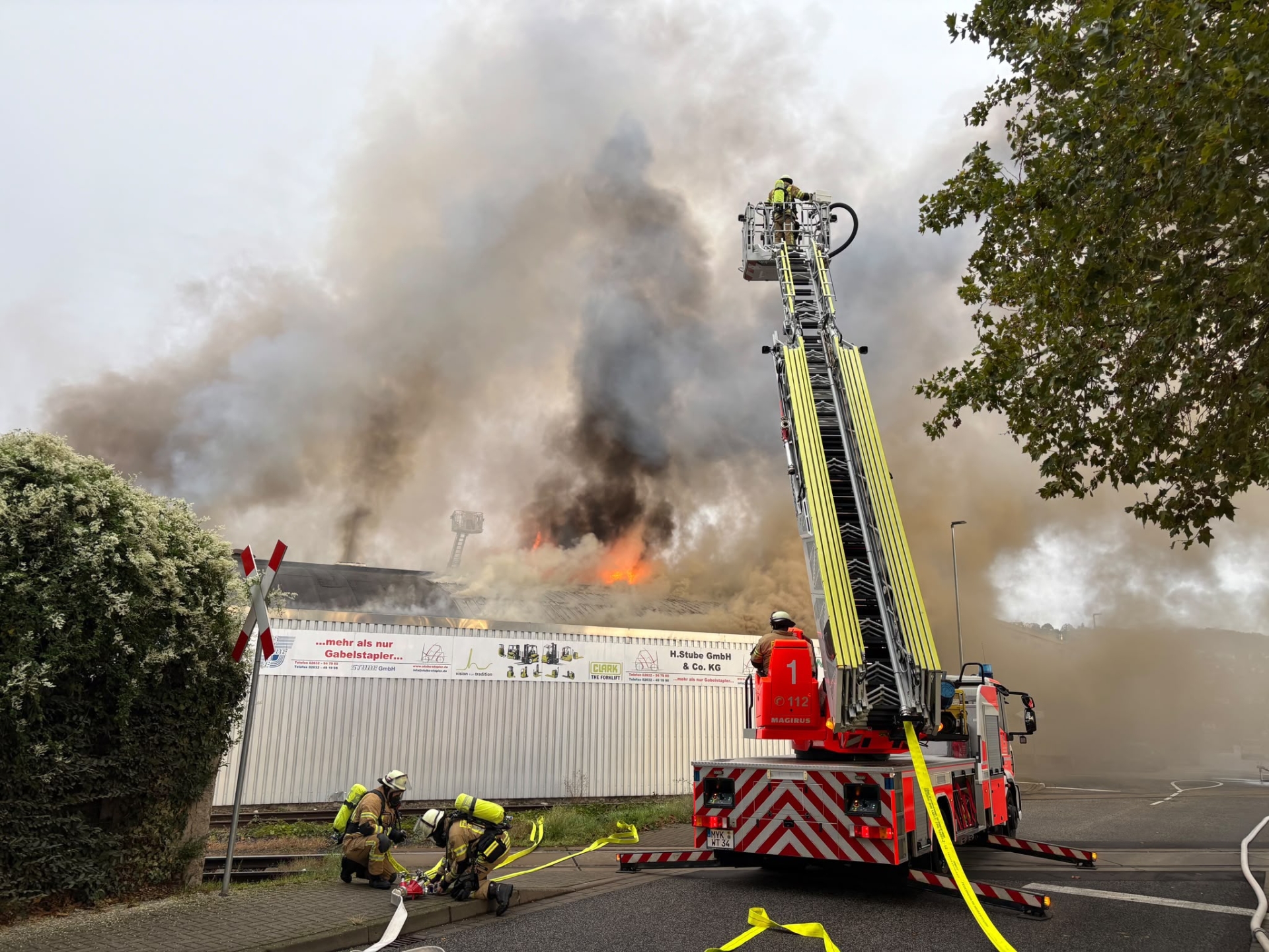 Hallenbrand in Andernach Foto 1 Vor einer brennenden Lagerhalle mit Flammen und Rauch stehen Feuerwehrleute auf der Drehleiter, während zwei weitere Feuerwehrleute am Boden mit Schläuchen beschäftigt sind.