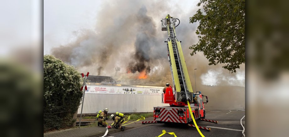 Hallenbrand in Andernach Foto 1 Vor einer brennenden Lagerhalle mit Flammen und Rauch stehen Feuerwehrleute auf der Drehleiter, während zwei weitere Feuerwehrleute am Boden mit Schläuchen beschäftigt sind.