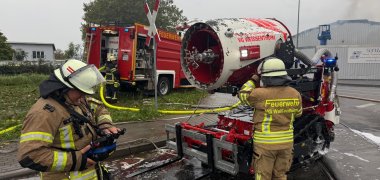 Hallenbrand in Andernach Foto 2 Zwei Feuerwehrleutesind am Löschroboter, der auf der Straße vor der Halle steht, beschäftigt. Die Straße ist nass vom Löschwasser. Auf einer Grünfläche neben der Straße steht ein Löschfahrzeug, davor ein Feuerwehrmann im Einsatz.