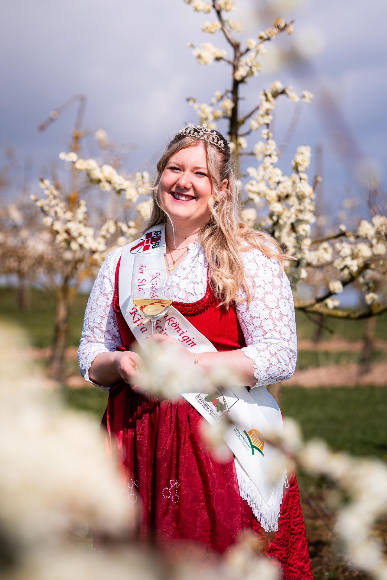 Kirschblütenkönigin Kira I. steht mit einem Glas Wein inmitten blühender Obstbäume. 