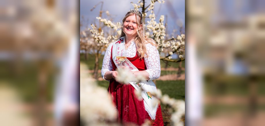 Kirschblütenkönigin Kira I. steht mit einem Glas Wein inmitten blühender Obstbäume. 