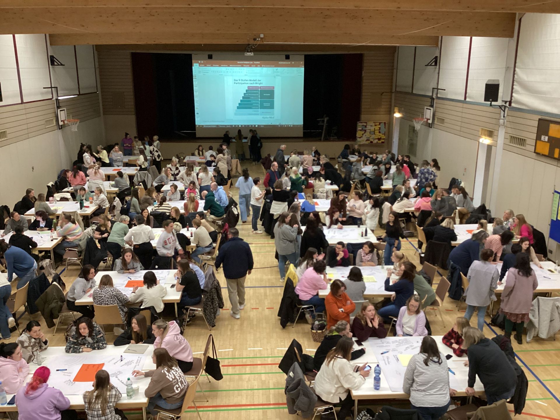 Von oben sieht man den Raum mit Publikum an Tischen, vorne auf der Bühne einen Bildschirm mit der Präsentation der Referentin.