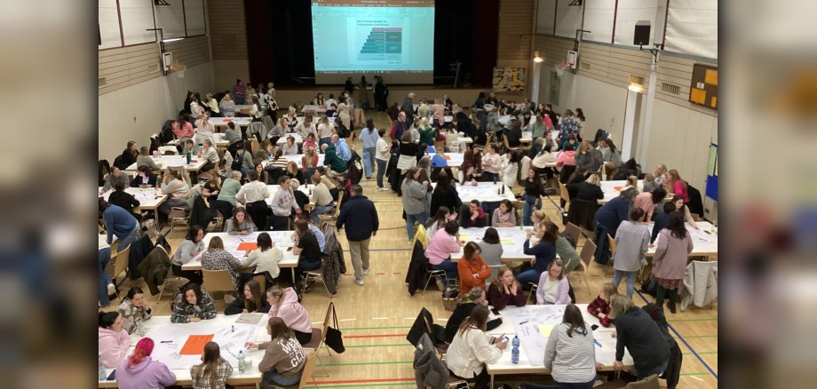 Von oben sieht man den Raum mit Publikum an Tischen, vorne auf der Bühne einen Bildschirm mit der Präsentation der Referentin.