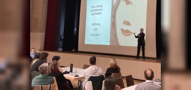 Die Referentin steht auf der Bühnen neben dem großen Bildschirm mit der Titelseite ihrer Präsentation.