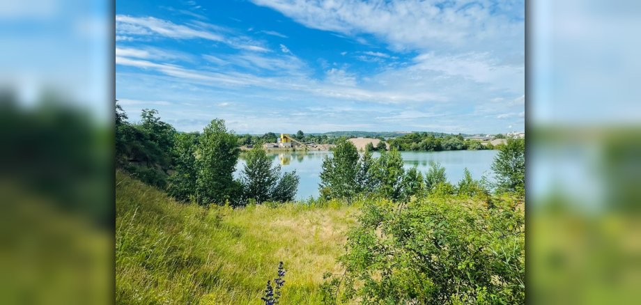 Unter sommerlich blauem Himmel ist der in die Landschaft eingebettete Kiesweiher zu sehen. 