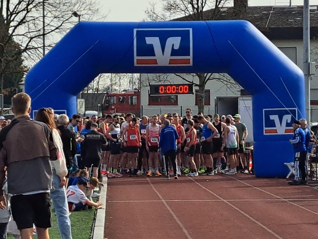 Unter einem blauen Start-Bogen stehen Läuferinnen und Läufer auf dem Sportplatz bereit zum Start des Firmenlaufs. 