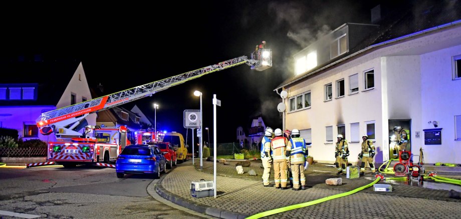 Vor dem Mehrfamilienhaus, in dem es brennt, steht ein Feuerwehrfahrzeug mit ausgefahrener Drehleiter und Feuerwehrkräften auf der Straße.