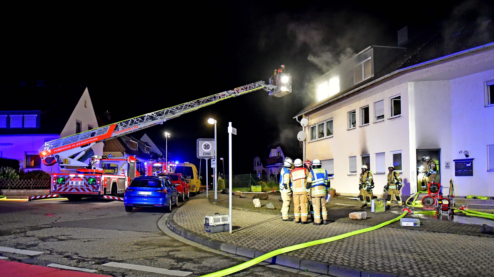 Wohnungsbrand Gesamtansicht Vor dem Mehrfamilienhaus, in dem es brennt, steht ein Feuerwehrfahrzeug mit ausgefahrener Drehleiter und Feuerwehrkräften auf der Straße.