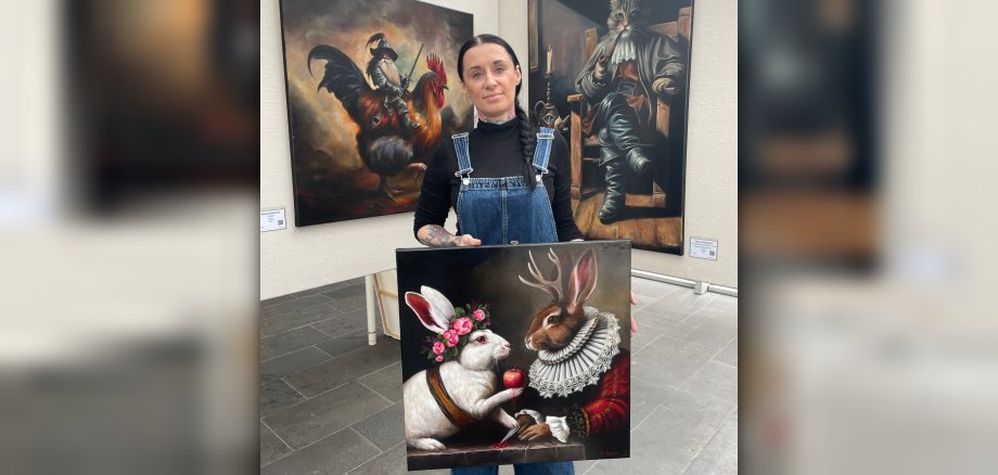 Ausstellung „Zwischen Traum & Wirklichkeit“ Künstlerin Anna Szóstak steht vor zwei Ausstellungswänden und hält eines ihrer Bilder in der Hand.