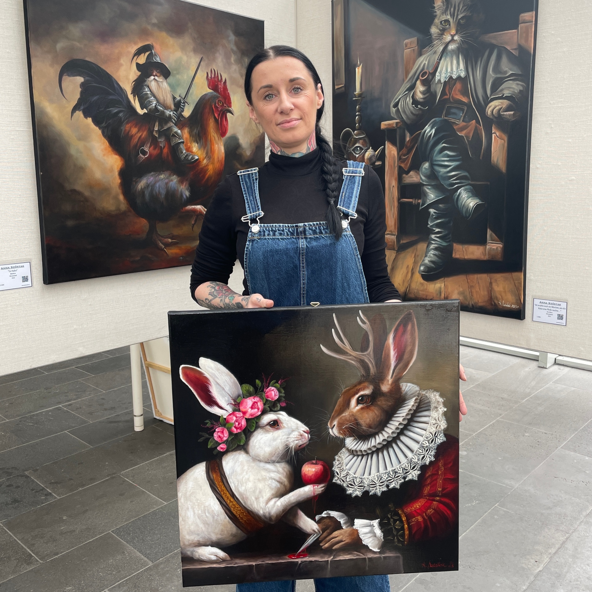 Ausstellung „Zwischen Traum & Wirklichkeit“ Künstlerin Anna Szóstak steht vor zwei Ausstellungswänden und hält eines ihrer Bilder in der Hand.
