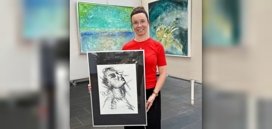 More than just colors“: Kunst im Foyer zeigt Werke, die Emotionen sichtbar machen Foto Künstlerin Miriam Lehnertz steht vor zwei Ausstellungswänden mit abstrakten Werken und hält eine gerahmte Skizze in den Händen.