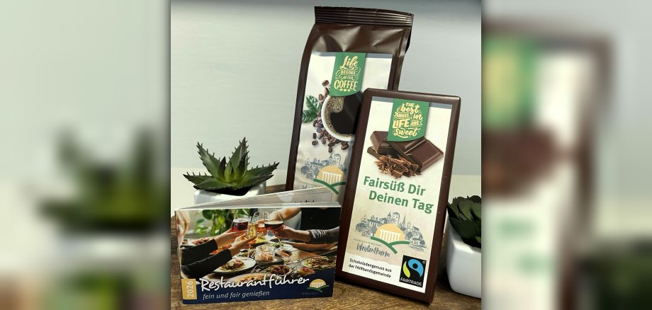 Das kleine Heftchen steht schräg vor Fairtrade-Kaffee und Fairtrade-Schokolade, daneben kleine Pflanzen.