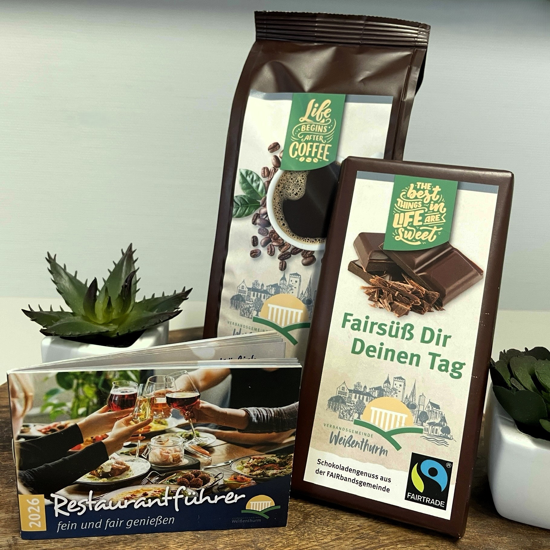 Restaurantführer 2026 Das kleine Heftchen steht schräg vor Fairtrade-Kaffee und Fairtrade-Schokolade, daneben kleine Pflanzen.