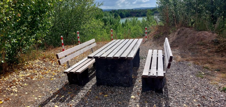 Ein fest installierter Tisch mit zwei Bänken steht in der Nähe  eines Abhangs, dahinter vier rot-weiß gestreifte Pfosten, im Hintergrund der Kiesweiher.