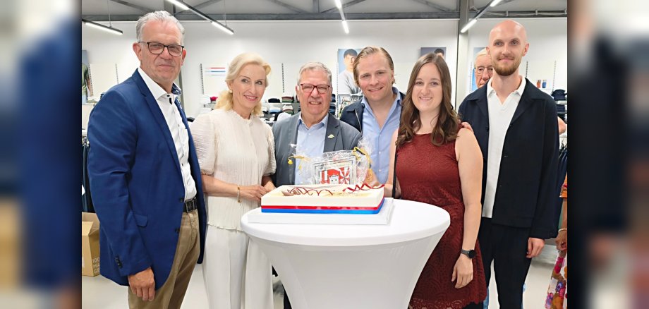 Eröffnung trigema Gruppenfoto Der Erste Beigeordnete Winfried F. Erbar und Wirtschaftsförderin Alina Kneip stehen mit der Unternehmerfamilie und Stadtbürgermeister Gerd Harner an einem Stehtisch, darauf eine Torte mit dem trigema-Logo.