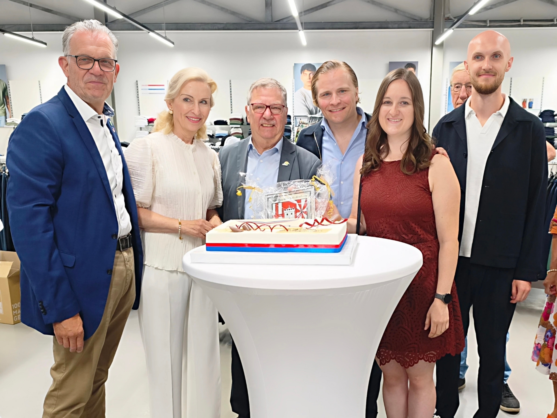Eröffnung trigema Gruppenfoto Der Erste Beigeordnete Winfried F. Erbar und Wirtschaftsförderin Alina Kneip stehen mit der Unternehmerfamilie und Stadtbürgermeister Gerd Harner an einem Stehtisch, darauf eine Torte mit dem trigema-Logo.