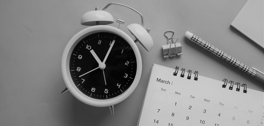 Auf einem grauen tisch liegt eine graue Uhr mit eine Kalender wo Termine drauf geschrieben werden können. 