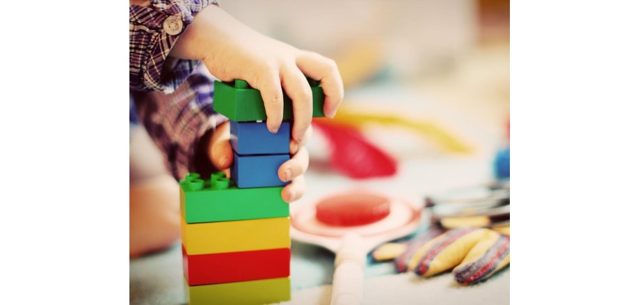 Im Vordergrund sind Kinderhände zu sehen, die mit großen Bausteinen einen Turm bauen. Im Hintergrund sind verschwommen eine Kelle, Handschuhe und weitere Bausteine zu sehen. 