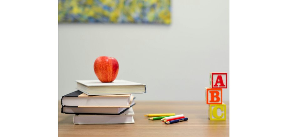 Auf einem Tisch liegen vier Bücher übereinander, darauf ein Apfel, daneben fünf Buntstifte und drei Würfel mit den Buchstaben A, B und C in rot, orange und gelb aufeinander. 