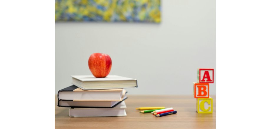 Auf einem Tisch liegen vier Bücher übereinander, darauf ein Apfel, daneben fünf Buntstifte und drei Würfel mit den Buchstaben A, B und C in rot, orange und gelb aufeinander. 