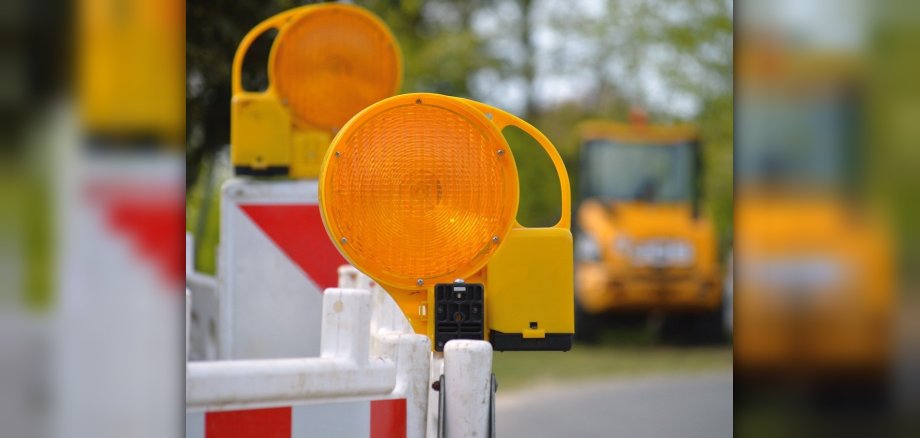 In Nahaufnahme eine Straßenabsperrung mit zwei orangefarbenen Leuchten zu sehen, im Hintergrund Bagger. 