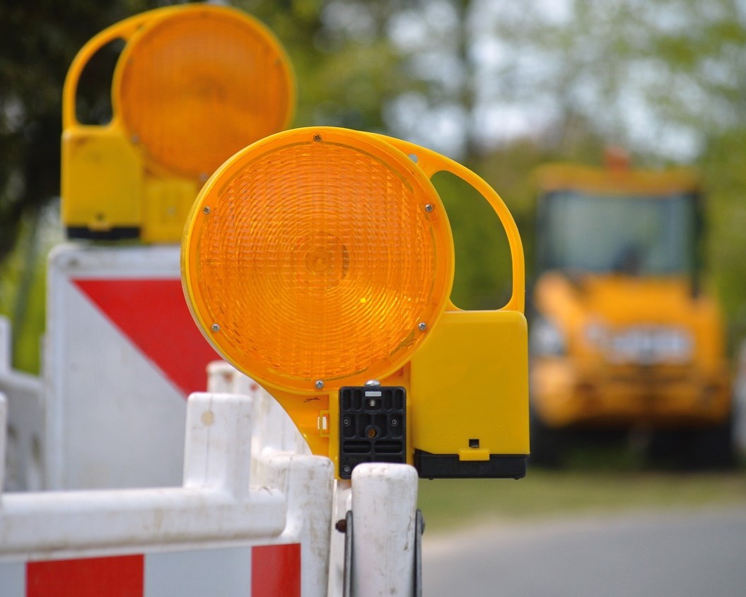 In Nahaufnahme eine Straßenabsperrung mit zwei orangefarbenen Leuchten zu sehen, im Hintergrund Bagger.