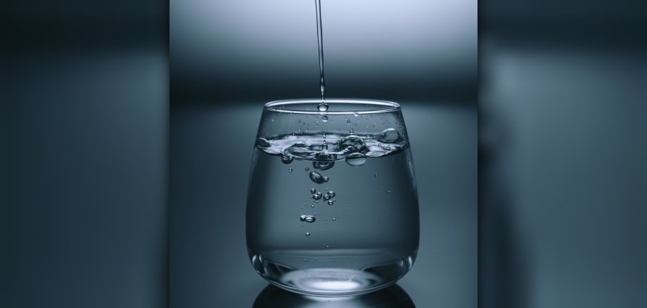 In ein fast volles Glas mit Wasser läuft von oben ein Strahl hinein und bildet Blasen unter der Oberfläche. 