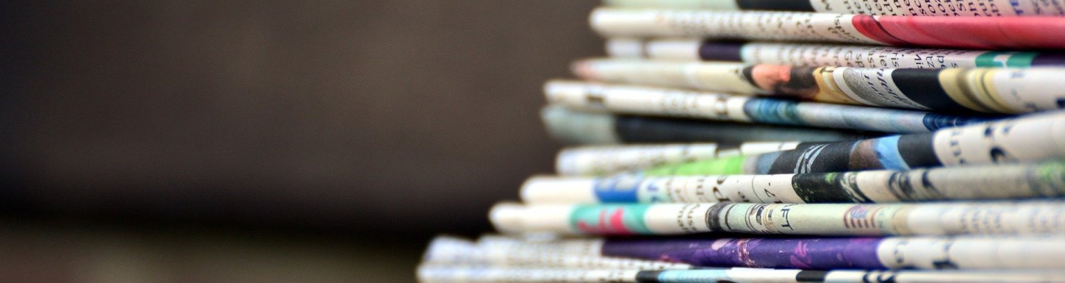 Ein Stapel Zeitungen liegt schräg rechts vorne auf einer Holzunterlage.