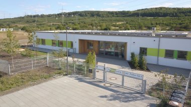 Die Kita ist weiß und an den Fenstern grün sie hat an dem Eingang eine schöne Holzoptik. 