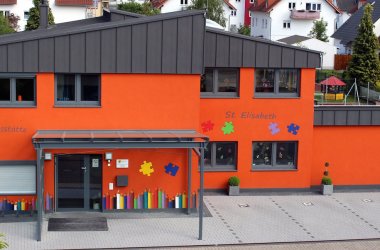 Die Kita ist orange das Dach ist schwarz der Eingang ist sehr schön bunt gestaltet 
